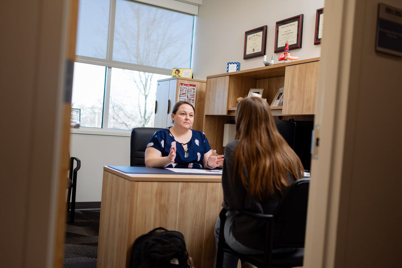 Faculty member at modern desk gestures while talking to student, back to camera, in brightly lit office.