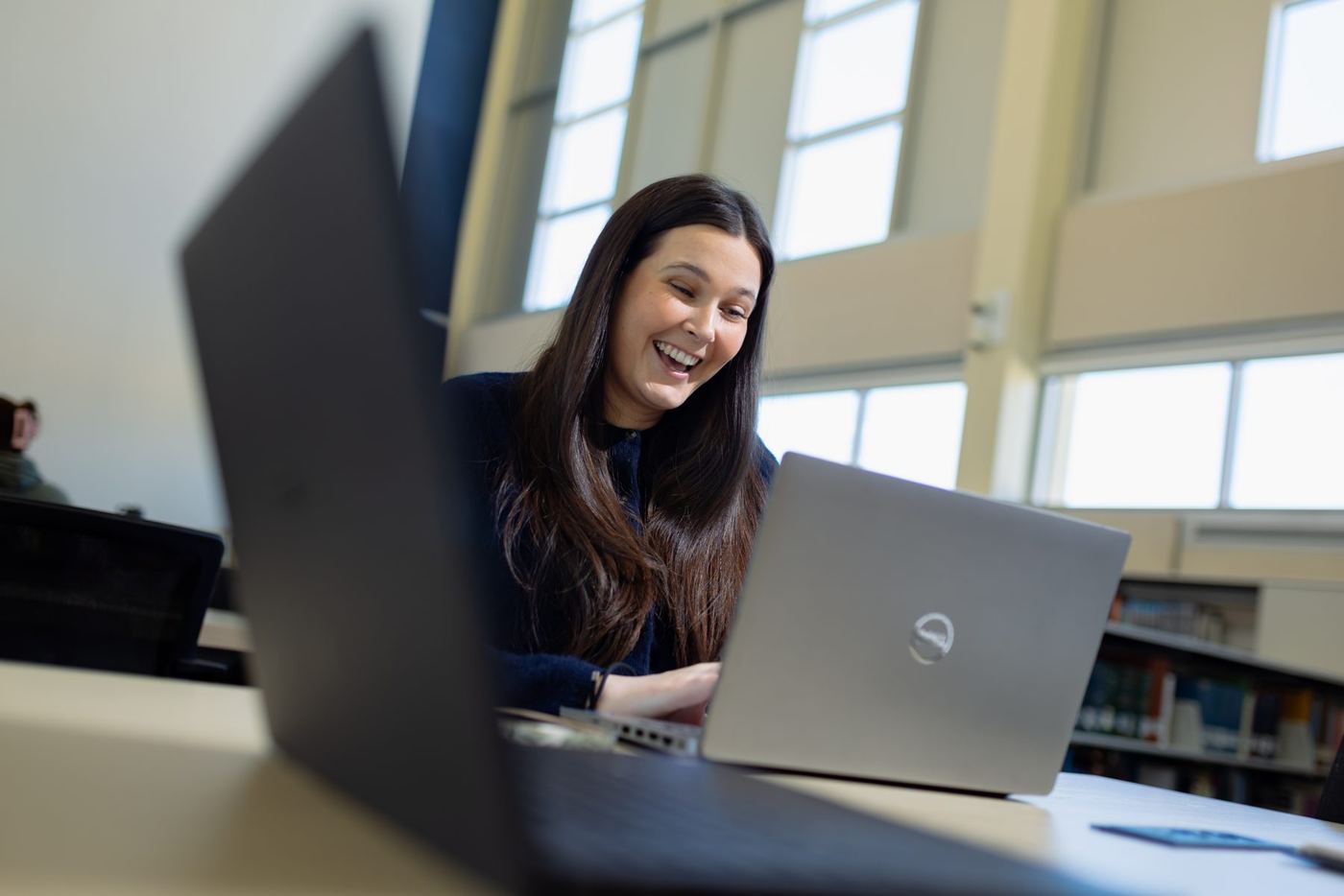 Smiling student with long dark hair typing on a silver laptop at a desk in a bright library.