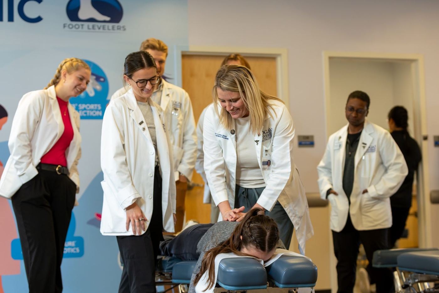 Chiropractic instructor demonstrates spinal adjustment to students in white lab coats.