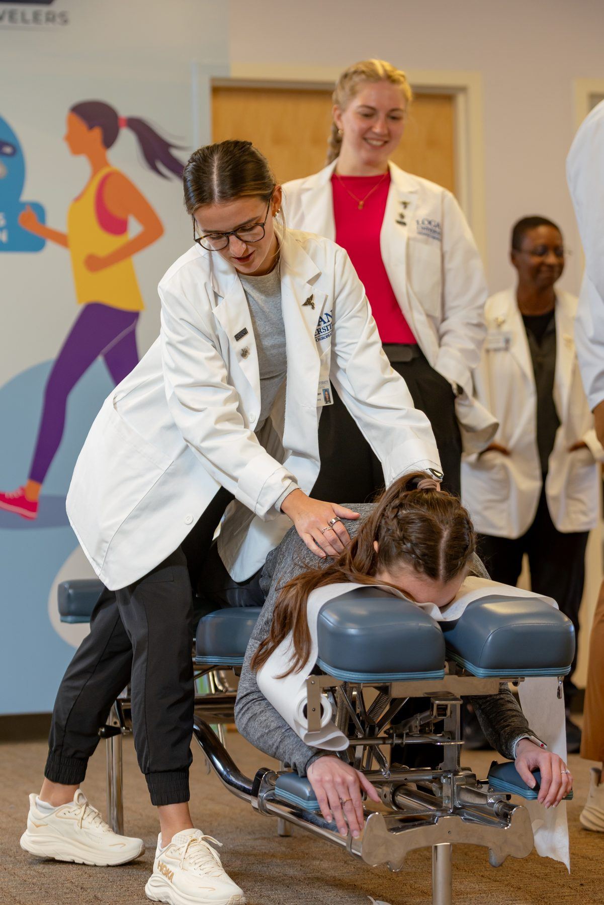 A student in a lab coat performs a chiropractic adjustment on a patient, with other students observing.