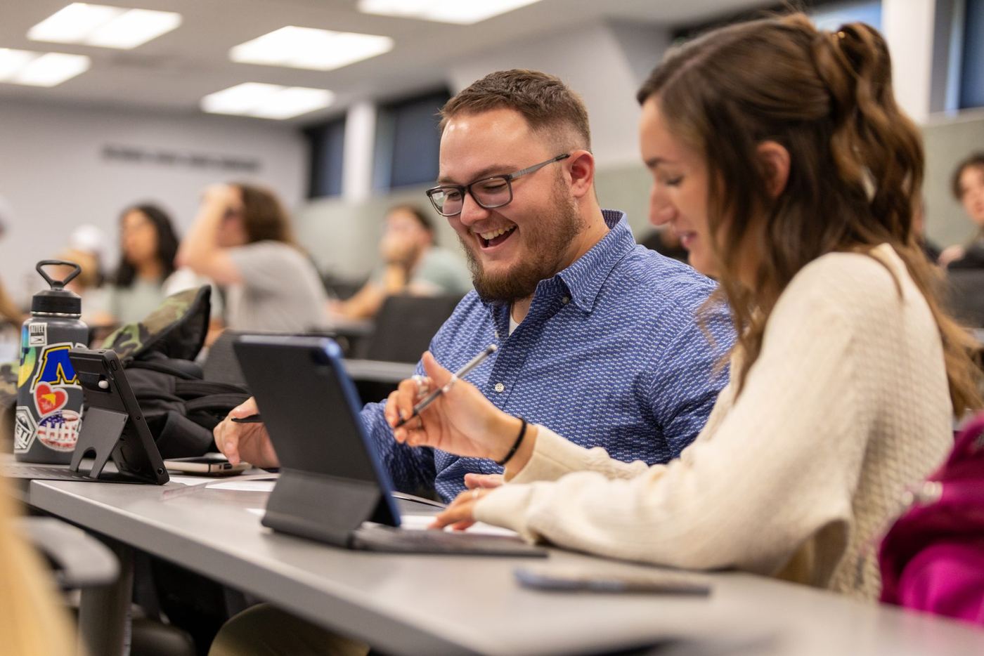 Man with glasses smiling at a tablet, next to a woman also using a tablet in a classroom.