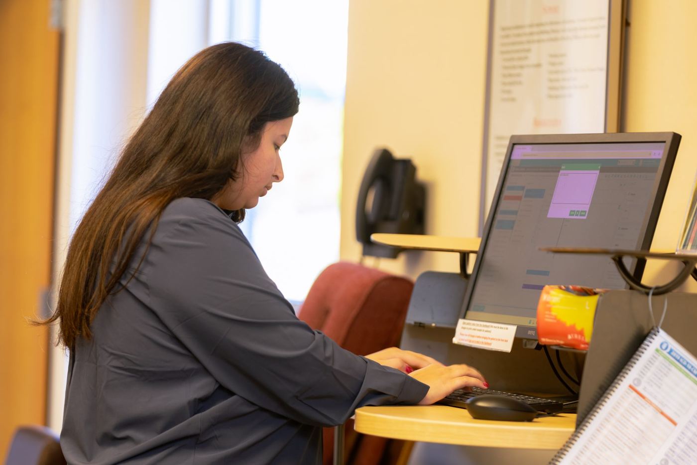 Woman with long hair typing on a computer, monitor shows a scheduling interface.