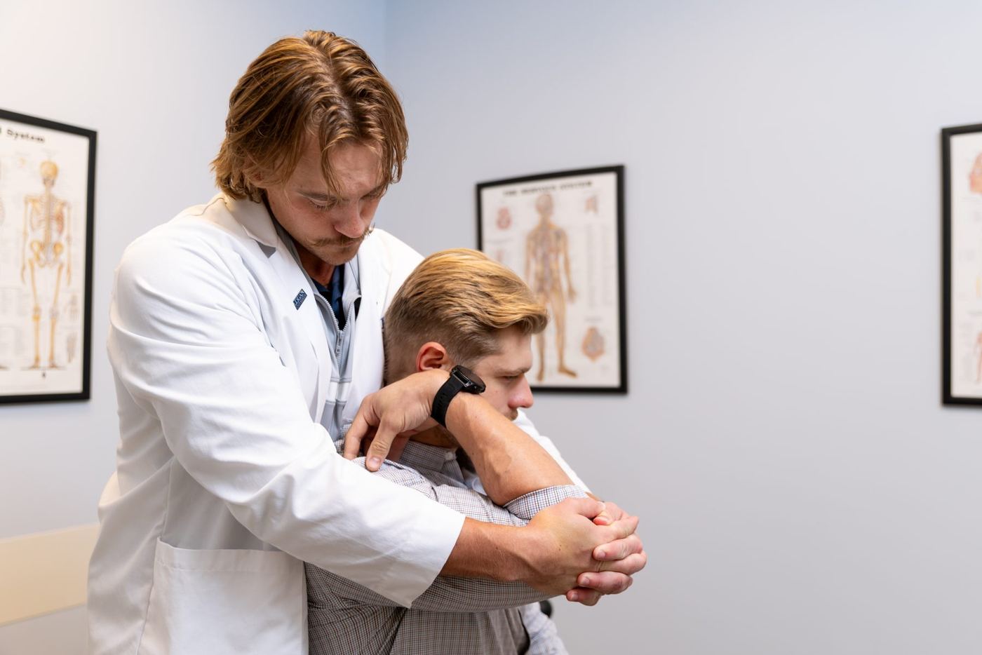 Male doctor in white lab coat performs a chiropractic adjustment on a patient's arm and shoulder.