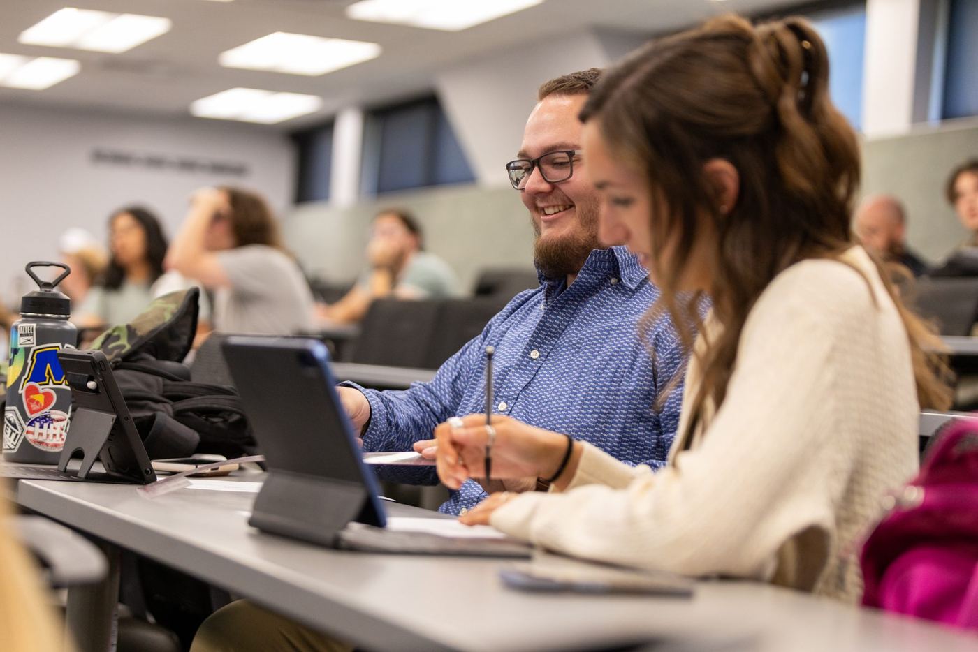 Two smiling Logan University students at desks with tablets and notes, in a bright classroom with others blurred.