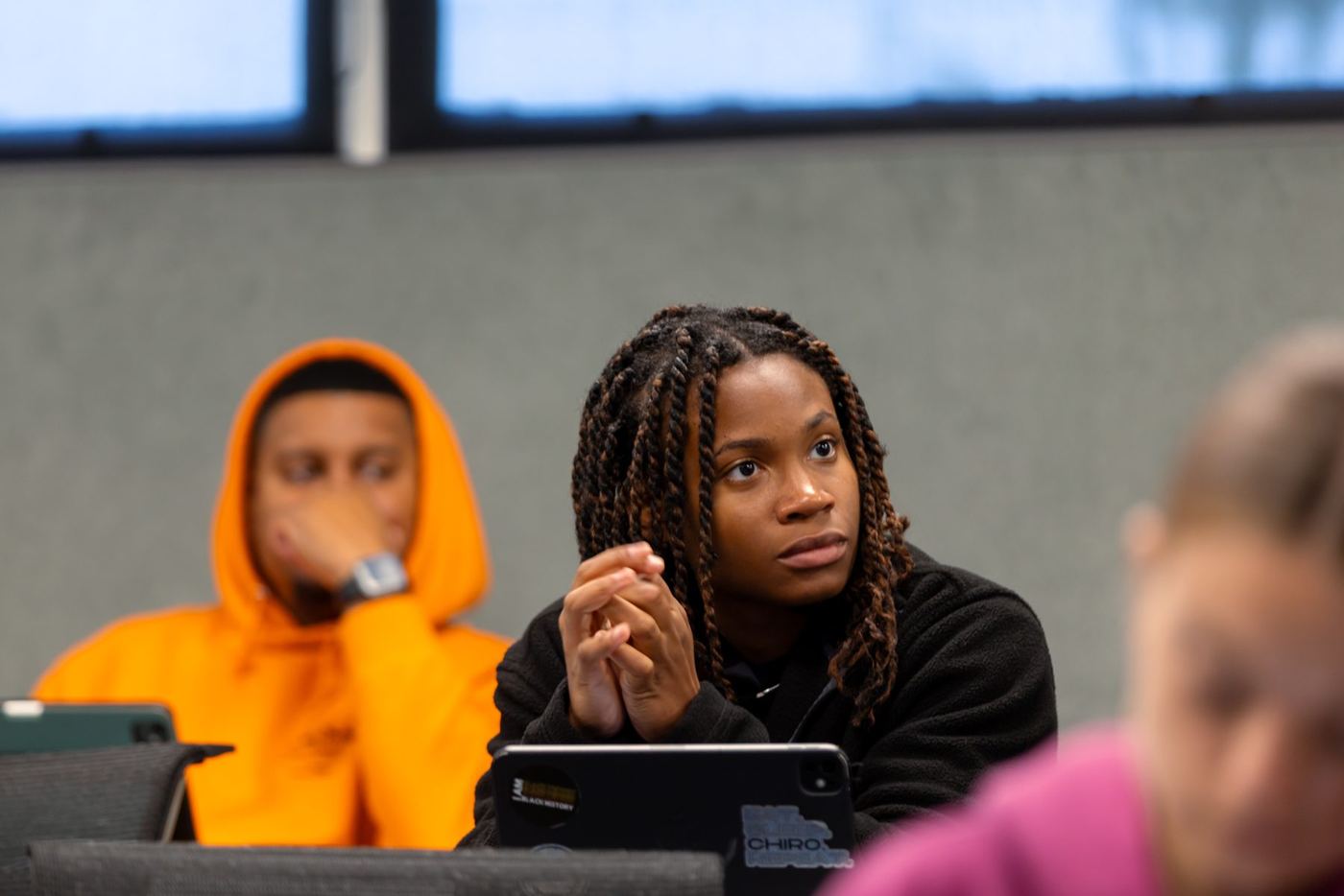 A young woman with braided hair intently watching off-camera in a classroom with other students.