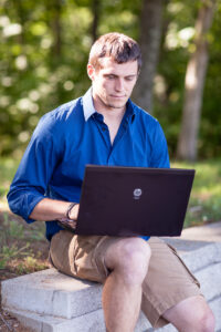 A man sits in a park and works on his laptop for his nutrition masters program