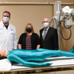 Three people stand behind a full-body radiology phantom lying on a table.