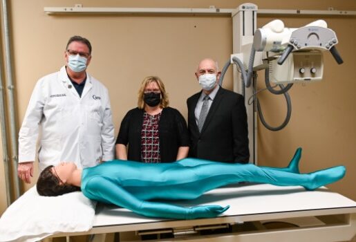 Three people stand behind a full-body radiology phantom lying on a table.