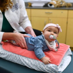 A Doctor of Chiropractic lays her hands on a baby's back with a gentle touch as the child looks toward the camera.